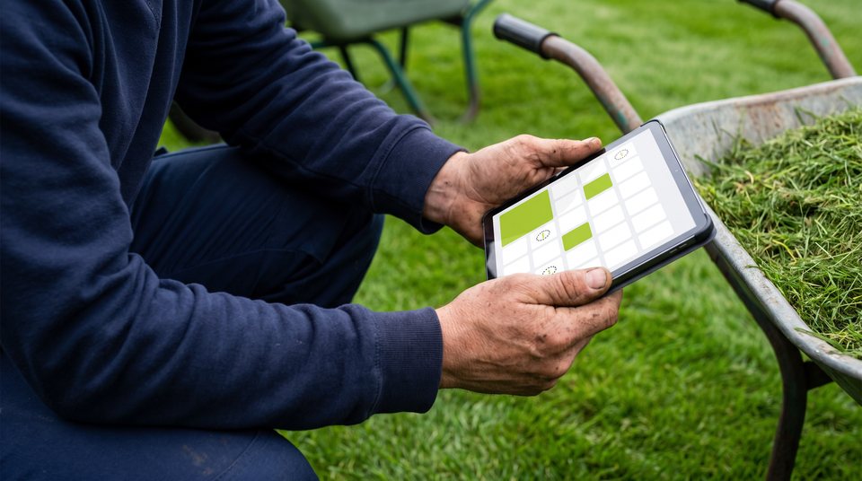 A landscaper kneeling on a lawn holds a tablet showing a weekly calendar grid with lime-green blocks, beside a wheelbarrow full of grass clippings